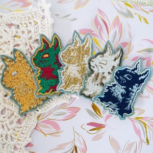 Peut inclure: Cinq écussons brodés en forme de lapin. Les couleurs incluent l'or, le vert avec des accents rouges, le blanc avec des accents marron et le bleu marine. Chaque écusson a une bordure bleu clair.