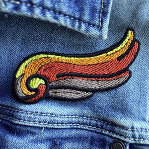 Puede incluir: Parche bordado con forma de ala en tonos amarillo, naranja, rojo y gris, con un contorno negro. El parche está adherido a una chaqueta vaquera azul, mostrando la textura de la tela y el diseño del parche.