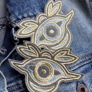 May include: Two embroidered patches with an eye-like design. The patches feature a gray base with intricate stitching in shades of blue, gold, and iridescent thread. The patches are placed on a blue denim jacket.