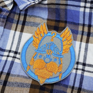 May include: Embroidered patch featuring a cartoon Viking with a blue helmet and orange beard, wings, and accents. The patch is gray with a blue border and a question mark on the helmet. The patch is on a blue, brown, and white plaid shirt.