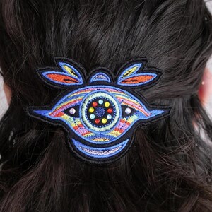 May include: A colorful embroidered hair clip featuring an eye design. The clip has a black base with blue, orange, yellow, and rainbow-colored embroidery. The eye has a central circle with colorful gems and is set against dark hair.