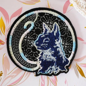 May include: Embroidered patch featuring a blue rabbit silhouette within a black circle. The rabbit is detailed with white and blue stitching, set against a floral pattern. The border has a light blue, white, and black design.