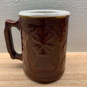以下が含まれることがあります： 白い縁取りと四つ葉のクローバーの浮き彫り模様が施された茶色の陶器製のマグカップ。マグカップには持ち手があります。