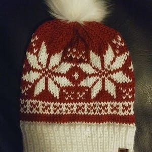 May include: A red and white knitted beanie with a white pom pom on top. The beanie has a snowflake pattern and a white cuff. The beanie has a brown leather tag with the text "K&K" on it.