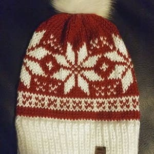 May include: Red and white knitted beanie with a white pom pom on top. The beanie has a snowflake pattern and a brown leather tag with the text "A.X.K."