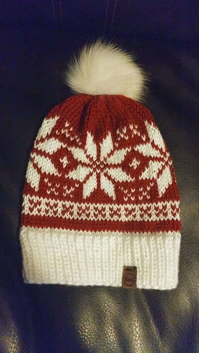 May include: Red and white knitted beanie with a white pom pom on top. The beanie has a snowflake pattern and a brown leather tag with the text "A.X.K."