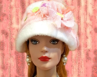 May include: A doll wearing a white and pink fuzzy bucket hat adorned with light green sequins and a pink fabric flower. The doll has blue eyes, red lips, and long reddish-brown hair, and is wearing large white pearl-like drop earrings.