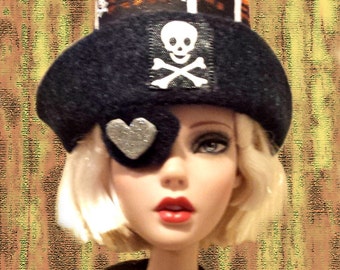 May include: A doll wearing a black pirate hat with a skull and crossbones patch and a silver heart-shaped eye patch. The hat has an orange and black patterned band. The doll has blonde hair and red lipstick.