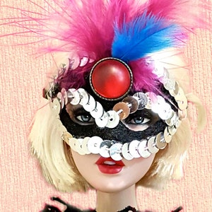 May include: A doll wearing a black sequined masquerade mask with a large red gem and pink and blue feathers. The doll has blonde hair, red lipstick, and a black lace top. The background is a light pink.