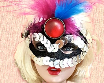 May include: A doll wearing a black sequined masquerade mask with a large red gem and pink and blue feathers. The doll has blonde hair, red lipstick, and a black lace top. The background is a light pink.