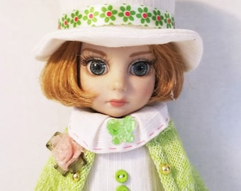 May include: A porcelain doll wearing a white dress with green and red floral trim, a green cardigan, and a white hat with a green and red floral ribbon. The doll has brown hair and blue eyes.