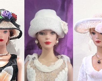 May include: Three dolls wearing different white hats. The doll on the left is wearing a black and white straw hat with a floral embellishment. The doll in the middle is wearing a white fluffy hat with a white flower. The doll on the right is wearing a white hat with a large white bow.