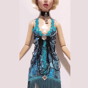 May include: A doll with blonde hair wearing a turquoise sequined dress with black beaded fringe and a matching headband featuring blue feathers and clear crystals. The outfit includes a black choker with a dangling crystal pendant.