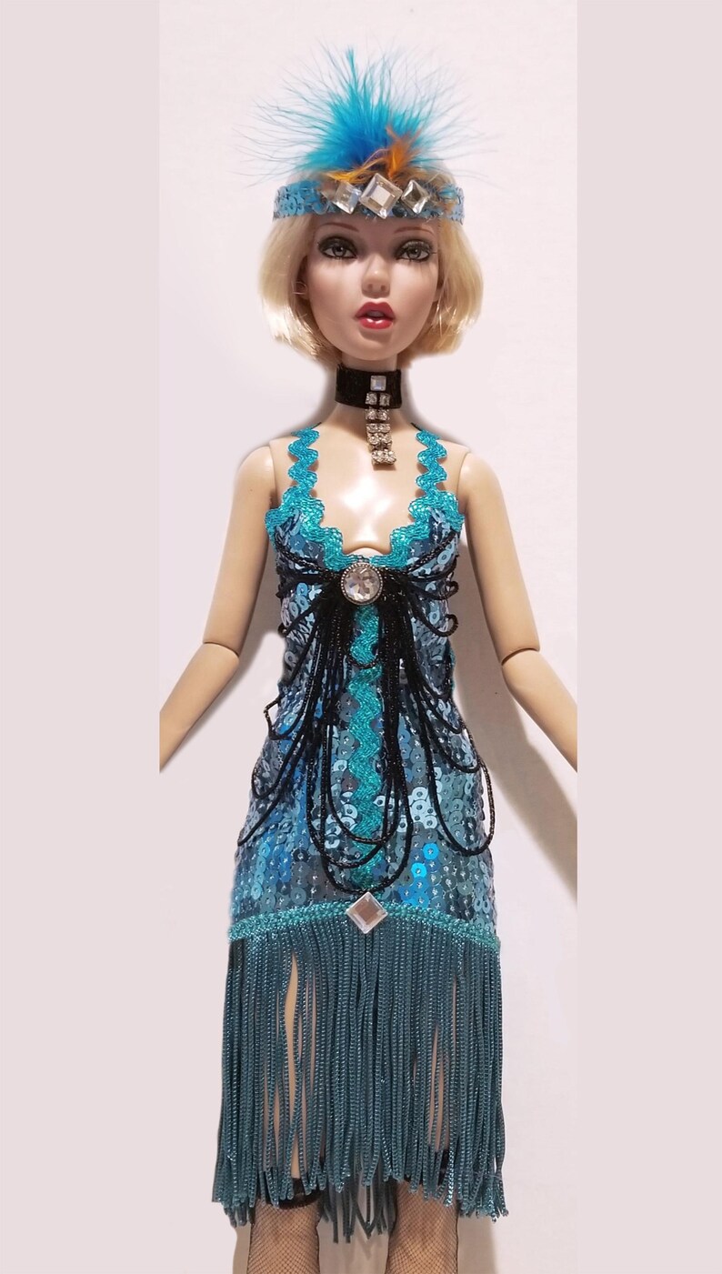 May include: A doll with blonde hair wearing a turquoise sequined dress with black beaded fringe and a matching headband featuring blue feathers and clear crystals. The outfit includes a black choker with a dangling crystal pendant.