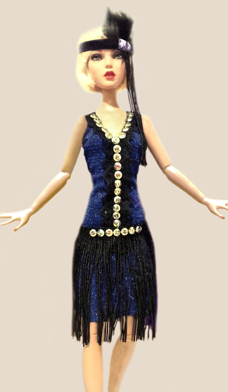 May include: A doll with blonde hair wearing a black headband with a feather and a dark blue dress. The dress features a V-neckline, a vertical row of silver sequins, black lace trim, and a black fringe skirt that hangs from the waist.