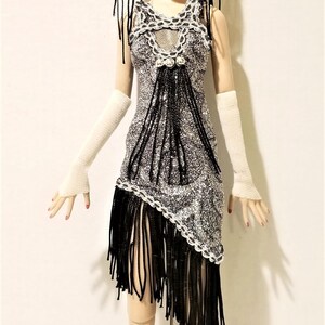 May include: A fashion doll with blonde hair wearing a black lace headpiece, a black choker, and a silver and black sleeveless dress with fringe detailing. The doll wears long white fingerless gloves, black fishnet stockings, and white heels.
