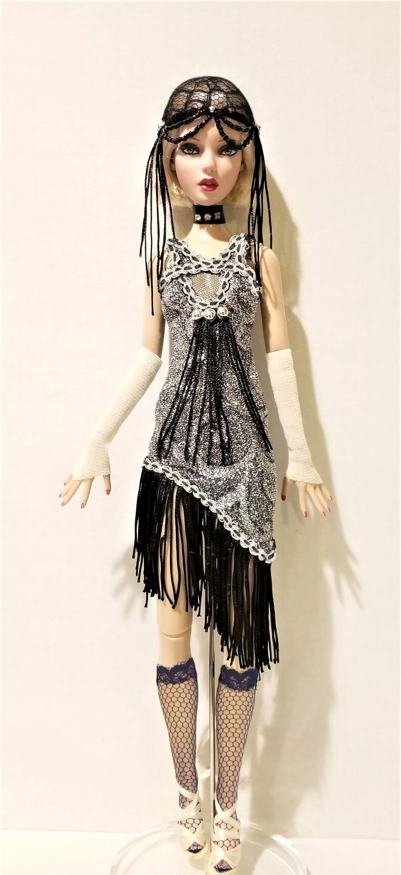 May include: A fashion doll with blonde hair wearing a black lace headpiece, a black choker, and a silver and black sleeveless dress with fringe detailing. The doll wears long white fingerless gloves, black fishnet stockings, and white heels.