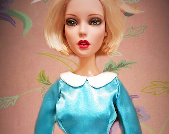 May include: A porcelain doll with blonde hair styled in a bob, wearing a light blue satin dress with a white collar and a white bow at the waist. The doll is posed against a pink background with a floral pattern.