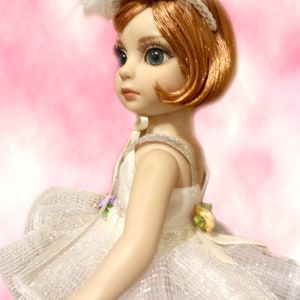 May include: A porcelain doll wearing a white tulle dress with a silver tiara and feather headband. The doll has red hair and blue eyes.