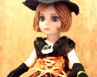 May include: A doll dressed in a Halloween witch costume. The doll wears a black and orange dress with a matching hat and holds a pumpkin-shaped bag. The hat has a glittery pumpkin design and a green pom-pom.