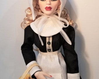 May include: A doll wearing a white bonnet and a black dress with a white apron. She is holding a basket with a red apple inside.