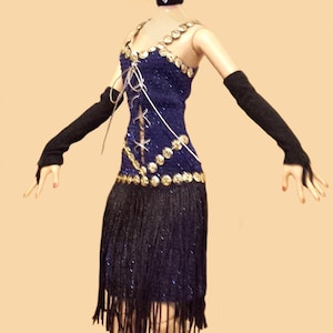 May include: A doll with blonde hair wearing a blue dress with gold sequins, a black fringe skirt, black opera gloves, a black choker, and a blue headband with red and black feathers. The doll wears white high-heeled sandals.