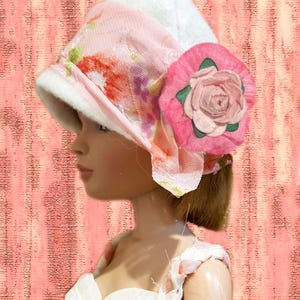K&ouml;nnte beinhalten: Ein wei&szlig;er Filzhut mit einem hellrosa Band mit Blumenmuster und einer gro&szlig;en rosa Stoffrose als Akzent an der Seite. Der Hut wird von einer Puppe mit braunem Haar getragen, kombiniert mit einem wei&szlig;en Kleid mit geflochtenen Tr&auml;gern.