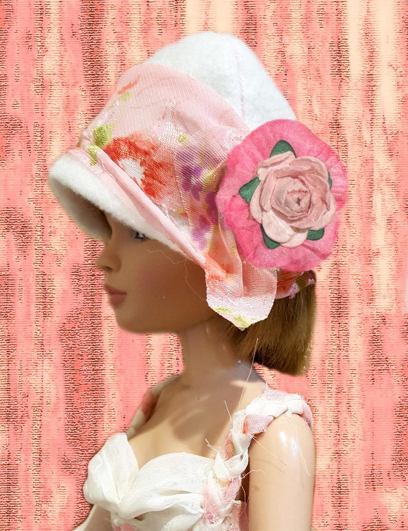 K&ouml;nnte beinhalten: Ein wei&szlig;er Filzhut mit einem hellrosa Band mit Blumenmuster und einer gro&szlig;en rosa Stoffrose als Akzent an der Seite. Der Hut wird von einer Puppe mit braunem Haar getragen, kombiniert mit einem wei&szlig;en Kleid mit geflochtenen Tr&auml;gern.