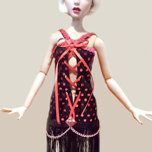May include: A doll with blonde hair wearing a black dress with red polka dots, a lace-up bodice, and a black fringe skirt. The doll wears a matching black and red headband with a red feather plume and a central square rhinestone accent.