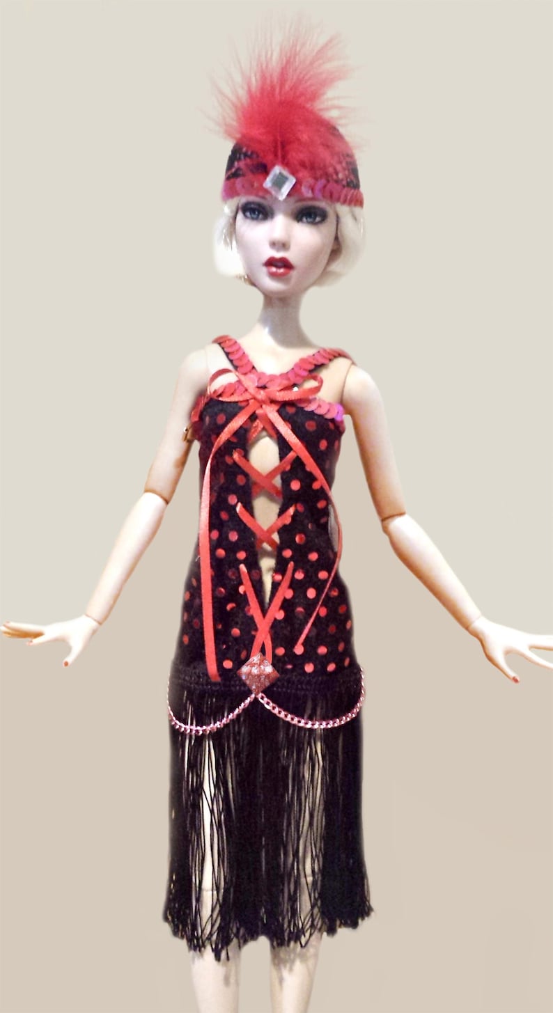 May include: A doll with blonde hair wearing a black dress with red polka dots, a lace-up bodice, and a black fringe skirt. The doll wears a matching black and red headband with a red feather plume and a central square rhinestone accent.