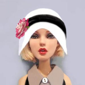 K&ouml;nnte beinhalten: Ein wei&szlig;er Glockenhut mit einem breiten schwarzen Band und einer rosa-wei&szlig;en Stoffblume an der Seite. Der Hut wird von einer Puppe mit blondem Haar, roten Lippen und einem grauen Hemd mit Kragen und einem wei&szlig;en Knopf getragen.