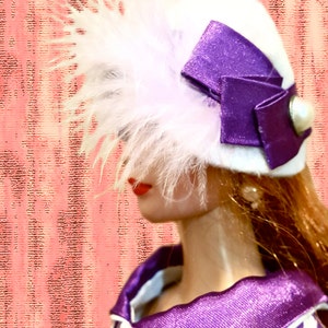 K&ouml;nnte beinhalten: Ein wei&szlig;er Hut mit einer gro&szlig;en violetten Satinschleife, einem wei&szlig;en Perlenakzent und hellrosa Federn. Der Hut wird zu einem wei&szlig;en Kleidungsst&uuml;ck mit violetten Punkten, einem violetten Kragen und einem passenden Knopf getragen.