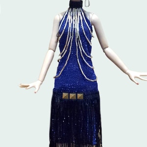 May include: A doll with blonde hair wearing a black lace headpiece with blue feathers, a blue halter dress featuring silver beaded strands, a black fringe skirt section, and three gold square studs at the waistline.