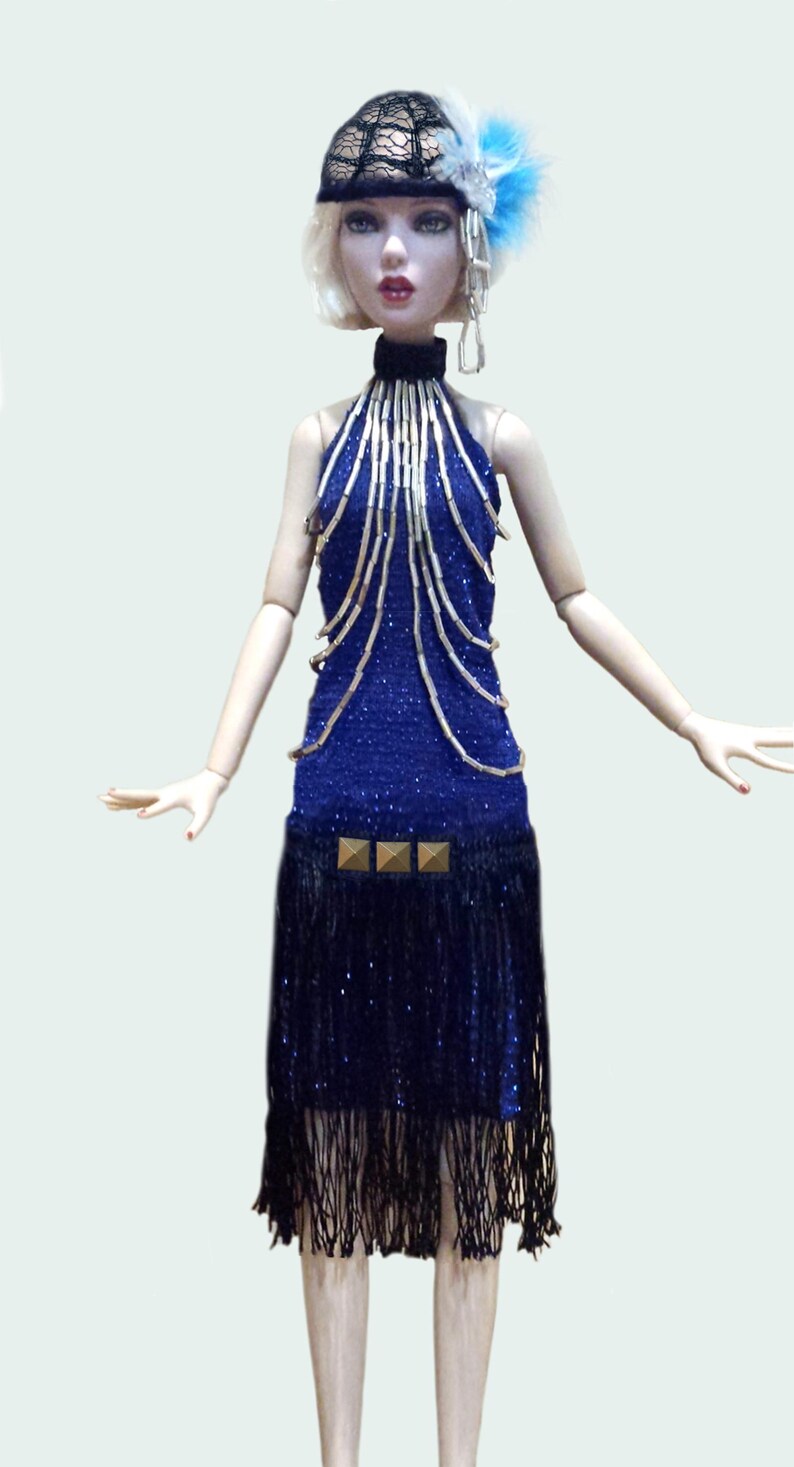 May include: A doll with blonde hair wearing a black lace headpiece with blue feathers, a blue halter dress featuring silver beaded strands, a black fringe skirt section, and three gold square studs at the waistline.
