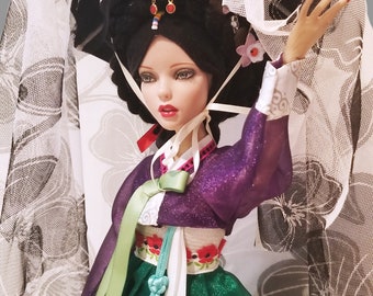 May include: A porcelain doll wearing a traditional Korean hanbok dress with a green skirt, purple top, and white sash. The doll has black hair and is wearing a white veil with floral designs.