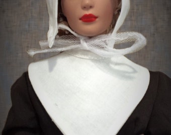 May include: A doll wearing a white bonnet and a white collar over a black dress. The doll has brown hair and red lips.