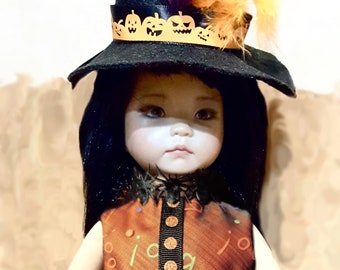May include: A Halloween-themed doll wearing a black witch hat with orange feathers and a pumpkin-themed band. The doll has black hair and is dressed in an orange and black dress with a spider collar. The doll is a Halloween decoration.