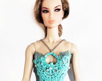 May include: A doll with brown hair wearing a turquoise crochet top with a silver chain necklace and a turquoise skirt.
