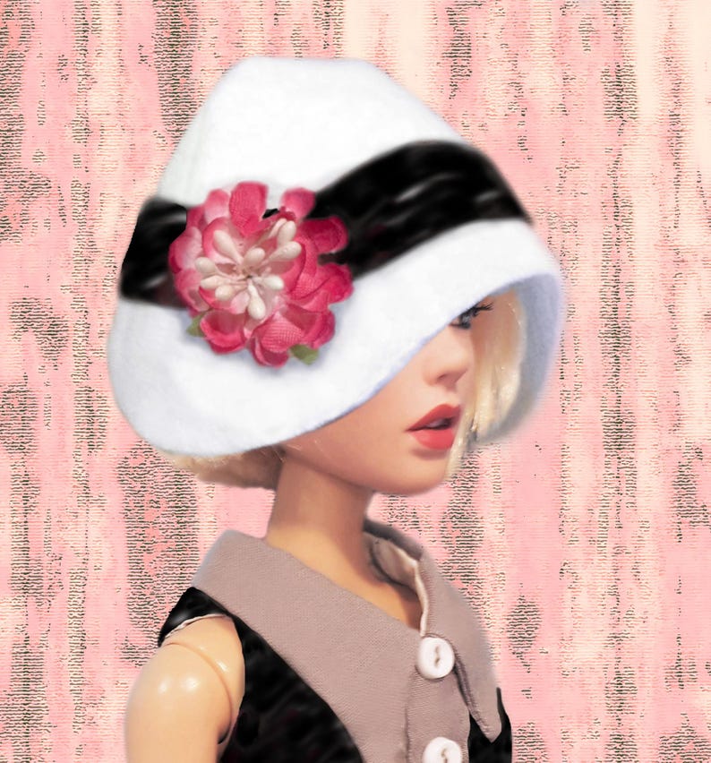 K&ouml;nnte beinhalten: Ein wei&szlig;er Glockenhut mit einem breiten schwarzen Band und einer gro&szlig;en, mehrlagigen rosa Stoffblume als Akzent. Der Hut wird von einer Puppe mit blondem Haar getragen, die ein schwarzes Kleidungsst&uuml;ck mit hellgrauem Kragen und wei&szlig;en Kn&ouml;pfen tr&auml;gt.