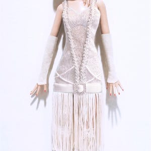 May include: A blonde fashion doll wearing a white sleeveless dress with a lace bodice, a drop waist with a square rhinestone buckle, and a long fringe skirt. The doll wears a white feathered headband, long white gloves, white fishnet stockings, and white open-toe heels.