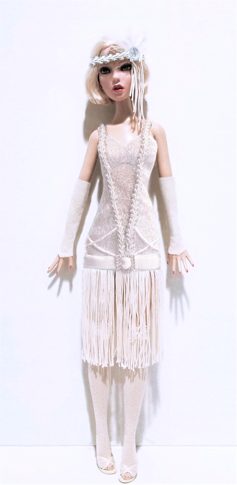 May include: A blonde fashion doll wearing a white sleeveless dress with a lace bodice, a drop waist with a square rhinestone buckle, and a long fringe skirt. The doll wears a white feathered headband, long white gloves, white fishnet stockings, and white open-toe heels.