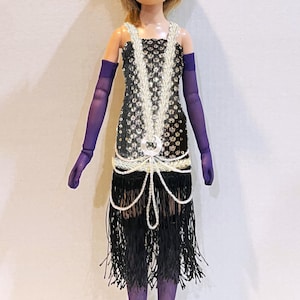 May include: A doll with blonde hair wearing a black sequined dress with white beaded fringe, purple opera-length gloves, purple tights, and a black feathered headpiece with a silver accent. The dress features a silver circular embellishment.