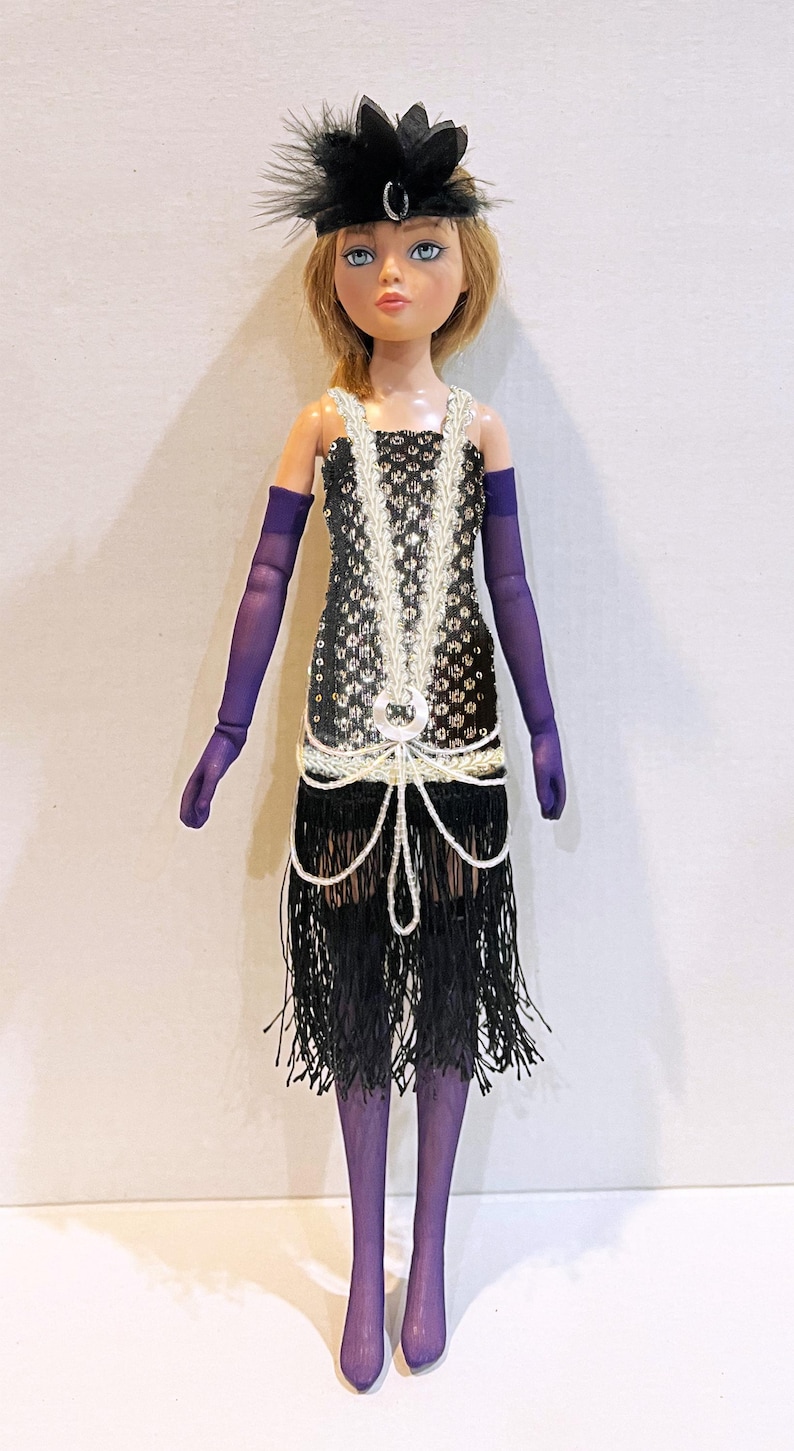 May include: A doll with blonde hair wearing a black sequined dress with white beaded fringe, purple opera-length gloves, purple tights, and a black feathered headpiece with a silver accent. The dress features a silver circular embellishment.