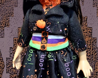 May include: A doll dressed in a Halloween costume. The doll wears a black jacket with a pumpkin button, a skirt with "boo!" text, and a pumpkin hat. The outfit features orange, purple, and green accents.