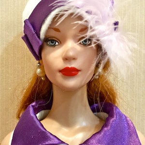 K&ouml;nnte beinhalten: Eine Puppe mit rotem Haar, blauen Augen und rotem Lippenstift tr&auml;gt einen wei&szlig;en Hut mit violettem Satinband und hellvioletten Federn. Die Puppe tr&auml;gt ein wei&szlig;es Hemd mit Kragen, violetten Punkten und einem einzelnen violetten Knopf.