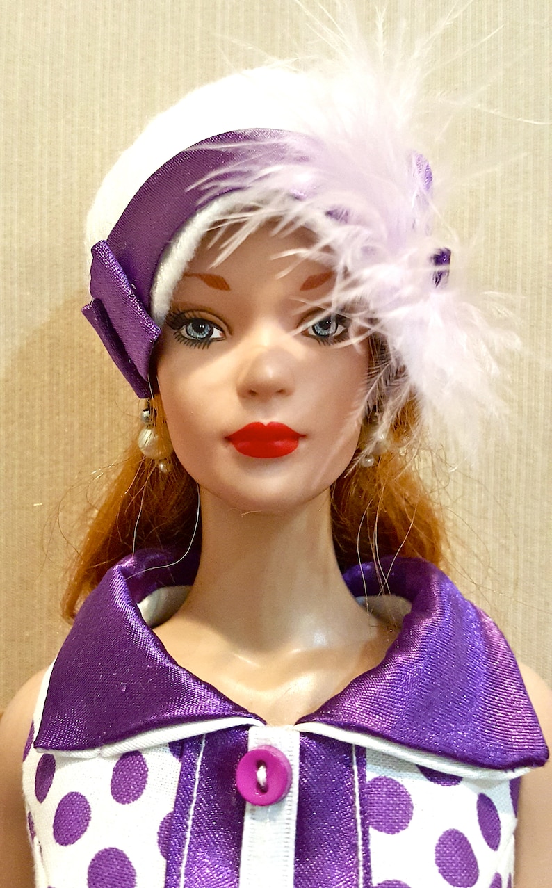 K&ouml;nnte beinhalten: Eine Puppe mit rotem Haar, blauen Augen und rotem Lippenstift tr&auml;gt einen wei&szlig;en Hut mit violettem Satinband und hellvioletten Federn. Die Puppe tr&auml;gt ein wei&szlig;es Hemd mit Kragen, violetten Punkten und einem einzelnen violetten Knopf.