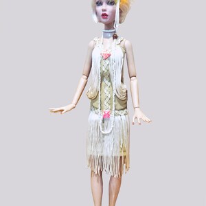 May include: A doll with short blonde hair wearing a yellow and orange feather headpiece, a white beaded headband, and a beige dress with white fringe layers. The outfit includes a long white beaded necklace, a choker, and beige strappy sandals.
