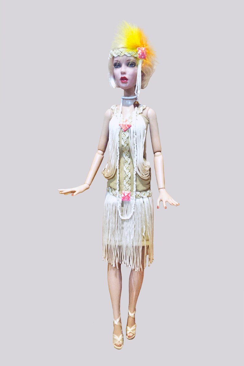 May include: A doll with short blonde hair wearing a yellow and orange feather headpiece, a white beaded headband, and a beige dress with white fringe layers. The outfit includes a long white beaded necklace, a choker, and beige strappy sandals.