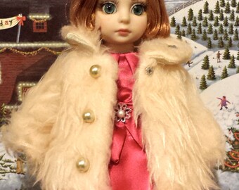 May include: A doll wearing a white faux fur coat with pearl buttons and a pink satin dress. The doll has red hair and blue eyes and is wearing a red bow with a pearl accent.