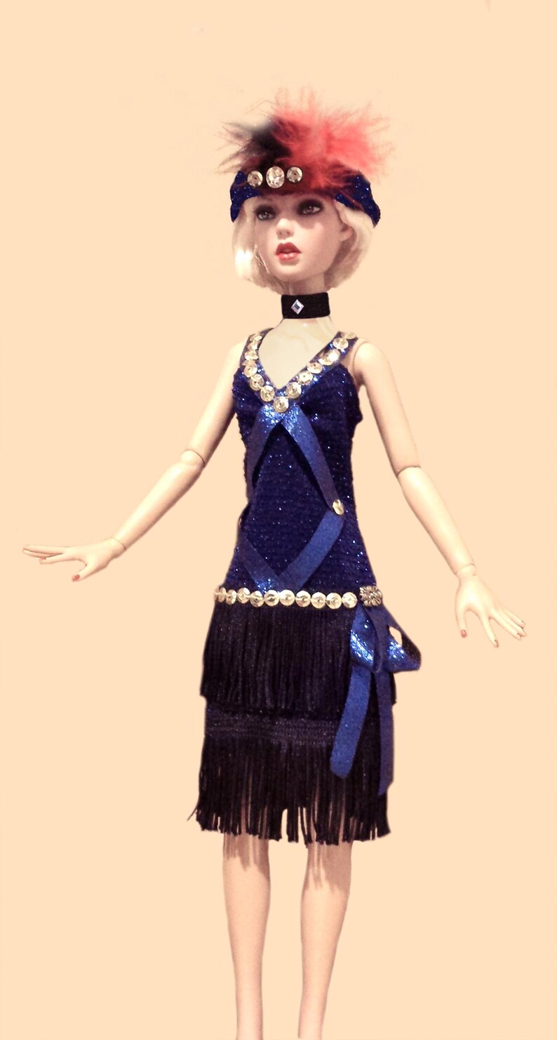 May include: A doll wearing a dark blue glittery dress with a V-neckline, silver sequin trim, and two tiers of black fringe. The doll has a black choker with a silver accent, a blue headband with silver sequins, and a red and black feather plume.